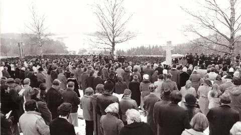 Ein schwarz weiß Foto. Eine Menschengruppe steht auf dem Friedhof in Grafeneck.  Im Hintergrund ein großes Steinkreuz