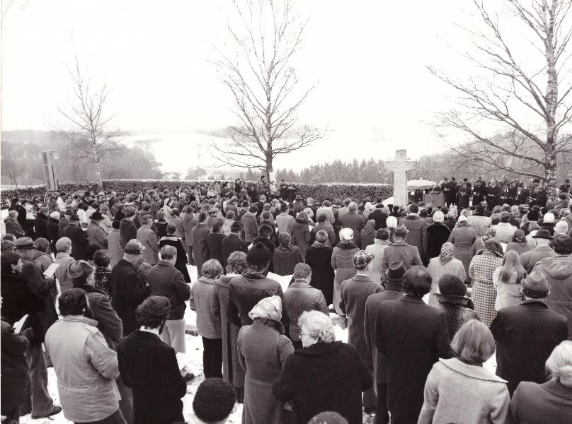 Ein schwarz weiß Foto. Eine Menschengruppe steht auf dem Friedhof in Grafeneck.  Im Hintergrund ein großes Steinkreuz